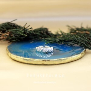 May include: A silver-colored ring with a twisted design and small, sparkling accents. The ring is displayed on a blue and gold-edged decorative plate, with greenery in the background. The Useful Bag handcrafted jewellery.