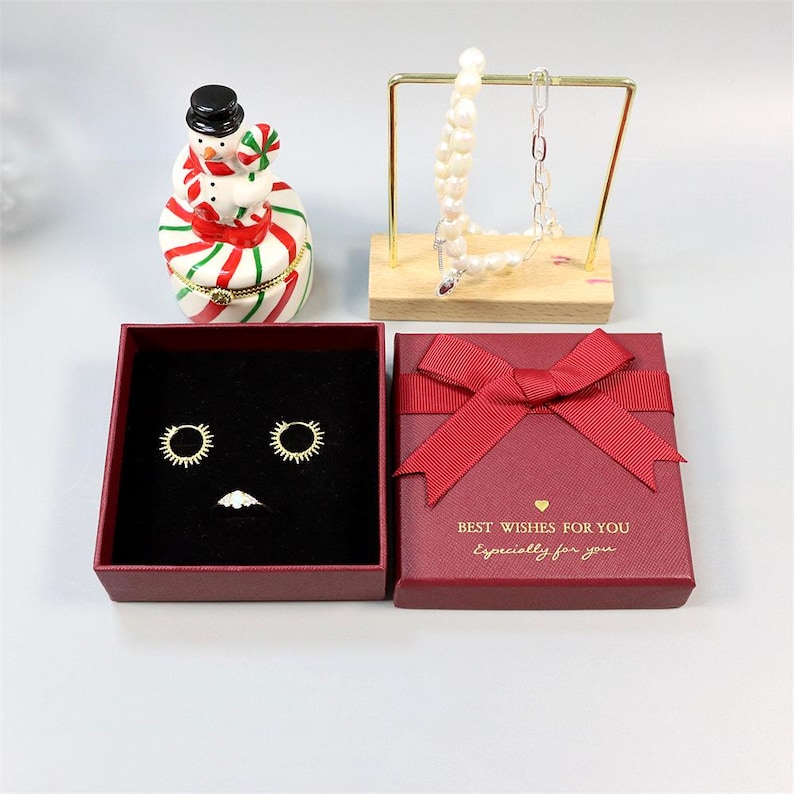 Op de afbeelding: Een assortiment sieraden en decoratieve items. Inclusief een rode geschenkdoos met strik, oorbellen, een ring, een sneeuwpopfiguur en een ketting op een displaystandaard. De geschenkdoos heeft de tekst "BEST WISHES FOR YOU Especially for you."