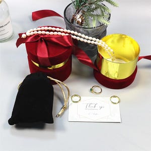 Op de afbeelding: Een rode fluwelen sieradendoos met een satijnen strik, een parelketting en een zwart fluwelen zakje. Ook een goudkleurige sieradendoos, gouden ringen en een bedankkaart.