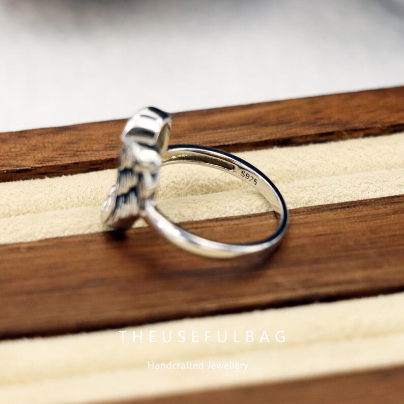 Op de afbeelding: Een zilveren ring met een gedetailleerd veermotief. De ring is gemarkeerd met "S925" op de band. De ring wordt gepresenteerd op een houten oppervlak. De tekst "THEUSEFULBAG" en "Handcrafted Jewellery" zijn zichtbaar.