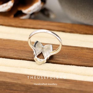 Op de afbeelding: Een zilveren ring met een vlindervleugelontwerp. De ring heeft een delicaat, opengewerkt patroon en is geplaatst op een houten oppervlak. De tekst "THEUSEFULBAG Handcrafted Jewellery" is onderaan zichtbaar.