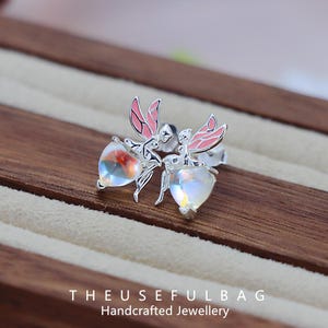 Peut inclure: Une paire de boucles d'oreilles fées en argent. Chaque boucle d'oreille représente une fée avec des ailes roses et une pierre précieuse irisée en forme de cœur. Le texte "THEUSEFULBAG Handcrafted Jewellery" est en bas.