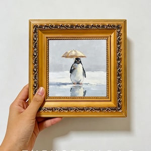 May include: A square painting in an ornate gold frame depicting a penguin standing on a snowy surface while holding a small beige umbrella. The artwork features a soft grey background and a reflection of the penguin on the icy ground below.