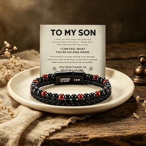 Peut inclure: Un bracelet de perles noires et rouges avec un bracelet en cuir tressé, présenté sur une assiette blanche avec une carte qui dit "TO MY SON". Le bracelet a un fermoir magnétique avec les mots "SHIELDED BY LOVE".