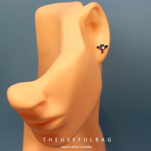 May include: A small, dark-colored earring with a circular, light-colored stone is displayed on a mannequin's ear. The earring has an ornate, wing-like design. The text "THEUSEFULBAG" and "Handcrafted Jeweller" are visible at the bottom of the image.