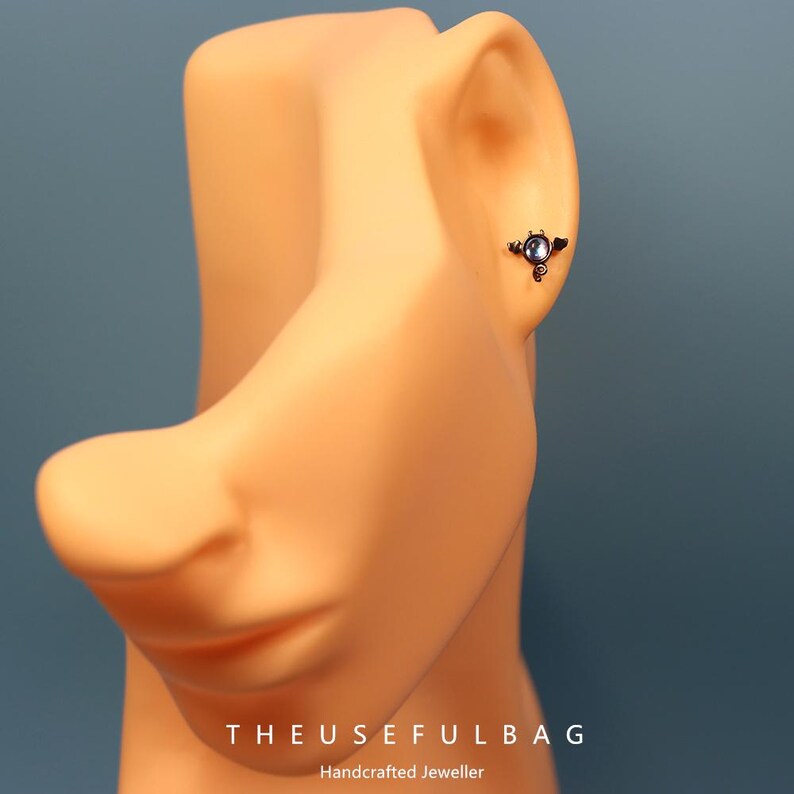May include: A small, dark-colored earring with a circular, light-colored stone is displayed on a mannequin's ear. The earring has an ornate, wing-like design. The text "THEUSEFULBAG" and "Handcrafted Jeweller" are visible at the bottom of the image.