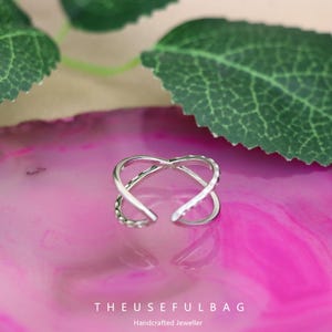 May include: A silver ring with an open design. The ring has an intertwined pattern, with textured and smooth sections. It is displayed on a pink surface, with green leaves in the background. The text "THEUSEFULBAG" and "Handcrafted Jeweller" are at the bottom.