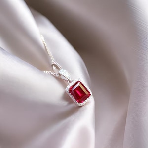 Puede incluir: Un collar de plata con un colgante de piedra roja rodeado de piedras transparentes más pequeñas. El colgante tiene forma cuadrada y el collar está sobre un fondo de tela sedosa.