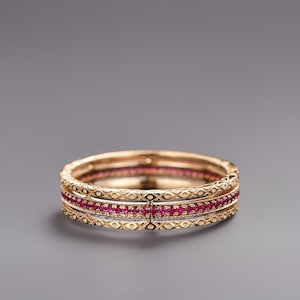 Puede incluir: Un conjunto de tres anillos dorados. El anillo central está adornado con pequeñas y vibrantes piedras preciosas rojas. Los anillos tienen un diseño intrincado y texturizado con un patrón repetido de pequeñas formas. Los anillos están apilados.