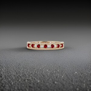 May include: A gold band ring with alternating red and white gemstones. The ring is set against a dark grey background. The gemstones are evenly spaced around the band, creating a symmetrical pattern.