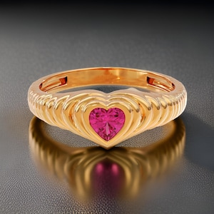 May include: A gold ring featuring a heart-shaped pink gemstone set in the centre. The band of the ring is textured with a series of raised lines.