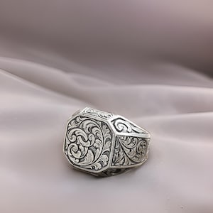 Puede incluir: Un anillo de plata con un intrincado diseño floral grabado. El anillo es de forma octogonal y tiene un acabado pulido.