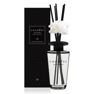Könnte beinhalten: Ein Cocod'or Black Edition Raumduft mit klarem Glasflakon, schwarzem Verschluss und schwarzen Stäbchen, die mit einer weißen Stoffblume verziert sind. Der Flakon trägt die Aufschrift Garden Lavender. Daneben steht eine hohe, schwarze Schachtel.