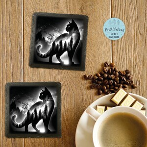 May include: Two black slate coasters with a black silhouette of a cat with a forest scene inside the cat. The cat is facing right and the forest scene is in the background. The coasters are on a wooden surface.