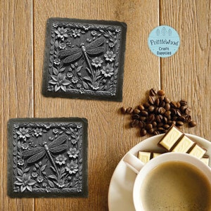 May include: Two square black slate coasters featuring a raised design of a dragonfly and flowers. The coasters are on a wooden surface with coffee beans and a cup of coffee.