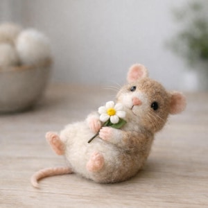 May include: A handmade felt mouse figurine in a relaxed pose, holding a white daisy with a yellow center. The mouse has tan and white fur, pink ears and paws, and black eyes. The background is blurred, with a wooden surface.