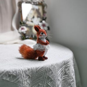 May include: A handcrafted felt squirrel figurine with a fluffy orange tail and a white lace skirt. The squirrel has a light brown body, dark brown feet, and a sweet expression. It stands on a white textured surface.