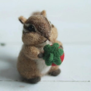 May include: A small, brown and white felt squirrel holding a green felt leaf and a red felt strawberry.