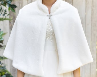 Puede incluir: Una capa de novia de piel sintética blanca con un cierre de botón decorativo. La capa se lleva sobre un vestido de novia blanco.