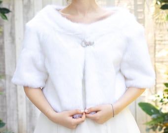 May include: A white faux fur bridal shrug with a small crystal brooch. The shrug is worn over a white wedding dress. The bride is standing in front of a green floral archway.