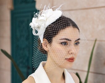 May include: A white fascinator hat featuring a structured base, delicate white feathers, and small pearl accents, paired with a white birdcage veil that covers the upper face. The headpiece is worn with a sleeveless white collared dress.