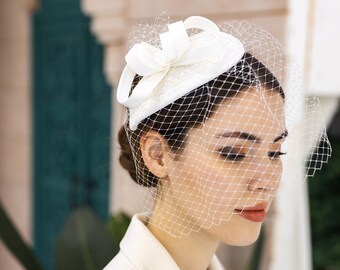 May include: A white bridal hat with a veil. The hat has a small, round brim and a bow on top. The veil is made of fine netting and covers the woman's face.