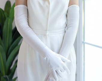 May include: A sleeveless white jumpsuit featuring a notched lapel collar and a button-front closure, paired with long, opaque white opera-length gloves that extend past the elbows, creating a coordinated monochromatic ensemble.