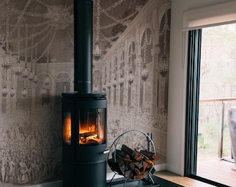 Può includere: Una stufa a legna nera con un fuoco che brucia all'interno. La stufa è di fronte a un muro con una carta da parati vintage che rappresenta una grande sala con archi e lampadari. Un porta legna in metallo nero si trova accanto alla stufa.