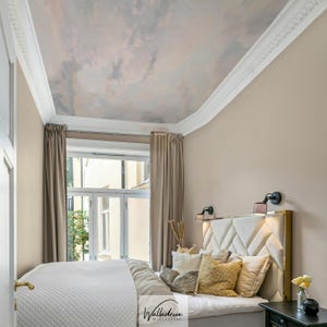 May include: A bedroom with a cloud-painted ceiling and beige walls. The bed has a white quilted coverlet, decorative pillows, and a gold-framed headboard. Beige curtains frame a window, and a small black nightstand is next to the bed.