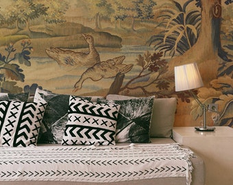 May include: A beige couch with a white throw blanket and black and white patterned pillows. The couch is in front of a tapestry wall mural depicting a forest scene with a river and two birds.