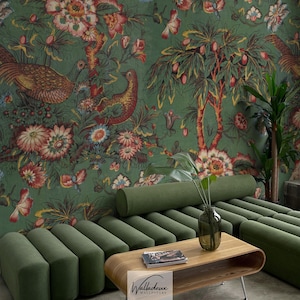 Op de afbeelding: Een woonkamer met een groene bank en een houten salontafel. De muur is bedekt met groen bloemenbehang met vogels en bloemen. Een vaas met planten staat op de salontafel. Het behang heeft de tekst "Walladeux Wallpapers".