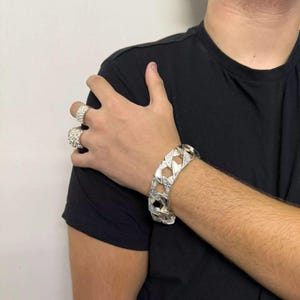 Puede incluir: Un brazalete de cadena de plata con un diseño de eslabones entrelazados gruesos. El brazalete se lleva en la muñeca de una persona que lleva una camiseta negra.