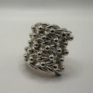Puede incluir: Un anillo de plata con un diseño texturizado que presenta un patrón tejido con pequeñas cuentas redondas.