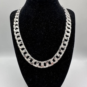 Peut inclure: Un collier en argent avec un design de chaîne à maillons plats. La chaîne est épaisse et a une finition brillante.