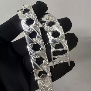 Op de afbeelding: Een zilveren armband met een gedetailleerd, sierlijk ontwerp. De ketting is dik en heeft een stoere, vierkante schakelstijl.
