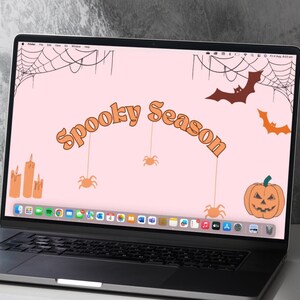 May include: A laptop computer with a pink background and a Halloween-themed desktop wallpaper. The wallpaper features the text "Spooky Season" in orange letters, a pumpkin with a jack-o'-lantern face, bats, spiders, and spiderwebs.