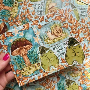 Könnte beinhalten: Eine Sammlung von Aufklebern mit skurrilen Illustrationen. Die Aufkleber zeigen Designs von Pilzen, Schnecken und Fröschen mit dem Text "Slow days are the Best". Die Aufkleber haben einen blauen Hintergrund mit orangefarbenen Blättern und Sternen.