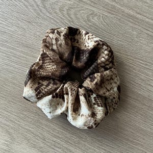 Peut inclure: Un chouchou avec un motif peau de serpent dans les tons marron, beige et crème. Le tissu est froncé et élastiqué, créant un accessoire texturé. Cet élastique à cheveux convient à diverses coiffures.