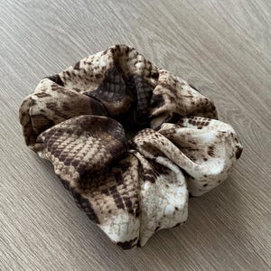 Op de afbeelding: Een scrunchie met een slangenprint in bruin, beige en cr&egrave;metinten. De stof is gerimpeld en gevouwen, wat een volumineuze, getextureerde uitstraling cre&euml;ert. Dit haaraccessoire is ontworpen om haar vast te houden en voegt een modieuze touch toe.
