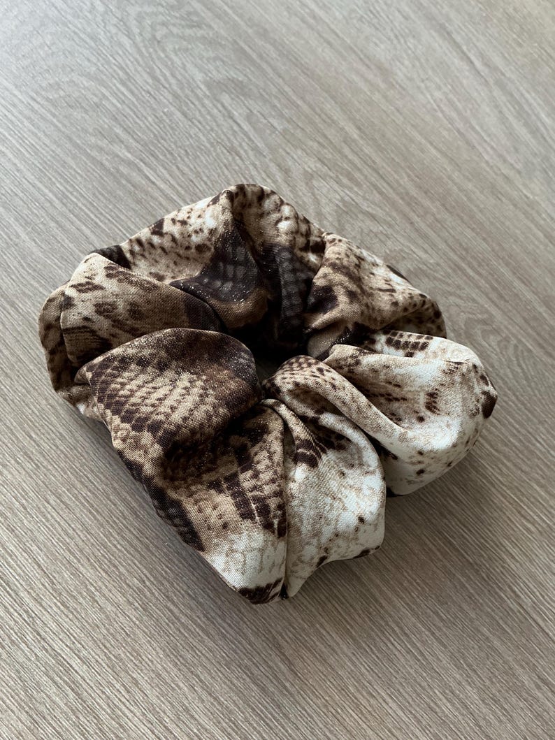 Op de afbeelding: Een scrunchie met een slangenprint in bruin, beige en cr&egrave;metinten. De stof is gerimpeld en gevouwen, wat een volumineuze, getextureerde uitstraling cre&euml;ert. Dit haaraccessoire is ontworpen om haar vast te houden en voegt een modieuze touch toe.