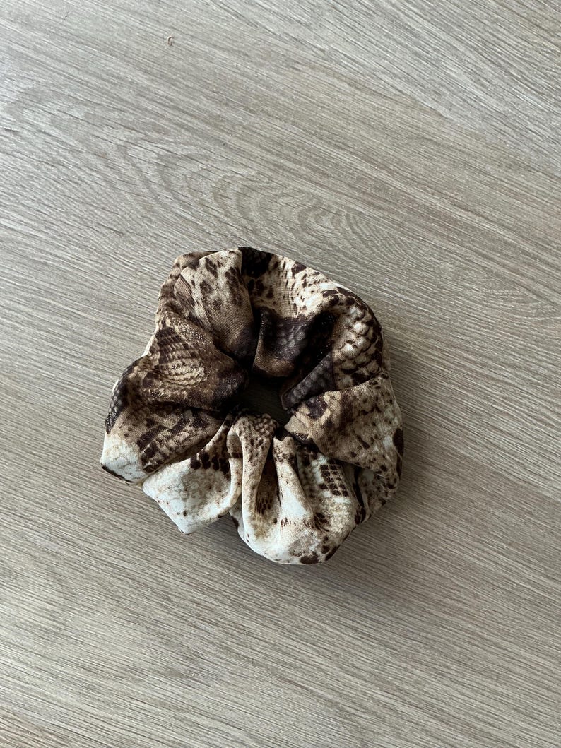 Op de afbeelding: Een scrunchie met een slangenprint in bruin, beige en cr&egrave;me. De stof is geplooid en elastisch, waardoor een accessoire met textuur ontstaat. Deze haarband is geschikt voor diverse kapsels.
