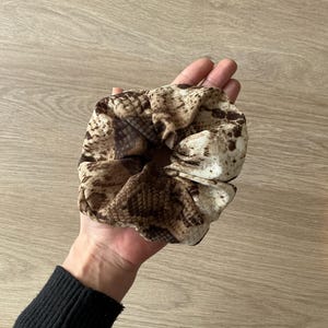 Op de afbeelding: Een scrunchie met een slangenprint in bruin- en beigetinten. Het haaraccessoire is gemaakt van een zachte, geplooide stof en wordt in een hand gehouden.
