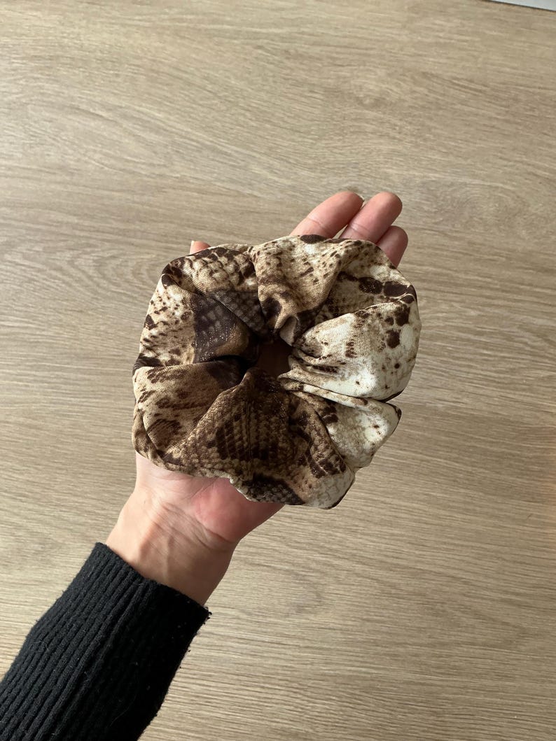 Op de afbeelding: Een scrunchie met een slangenprint in bruin- en beigetinten. Het haaraccessoire is gemaakt van een zachte, geplooide stof en wordt in een hand gehouden.