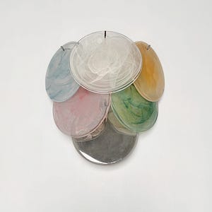 Könnte beinhalten: Eine Ansammlung von durchscheinenden, kreisförmigen Dekorationselementen in verschiedenen Farben. Die Farben umfassen Blau, Rosa, Grün, Gelb und eine silberfarbene Scheibe am Boden. Jede Scheibe hat ein einzigartiges Muster und ist mit kleinen Metallbefestigungen versehen.