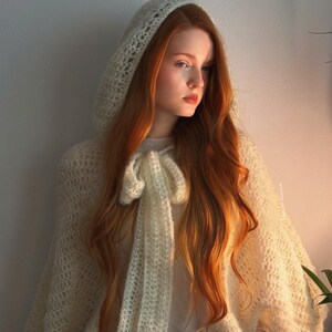 May include: A cream-colored, hooded crochet cape with a bow detail. The cape is worn over a white long-sleeved shirt. The model has long, red hair and is lit by warm sunlight.