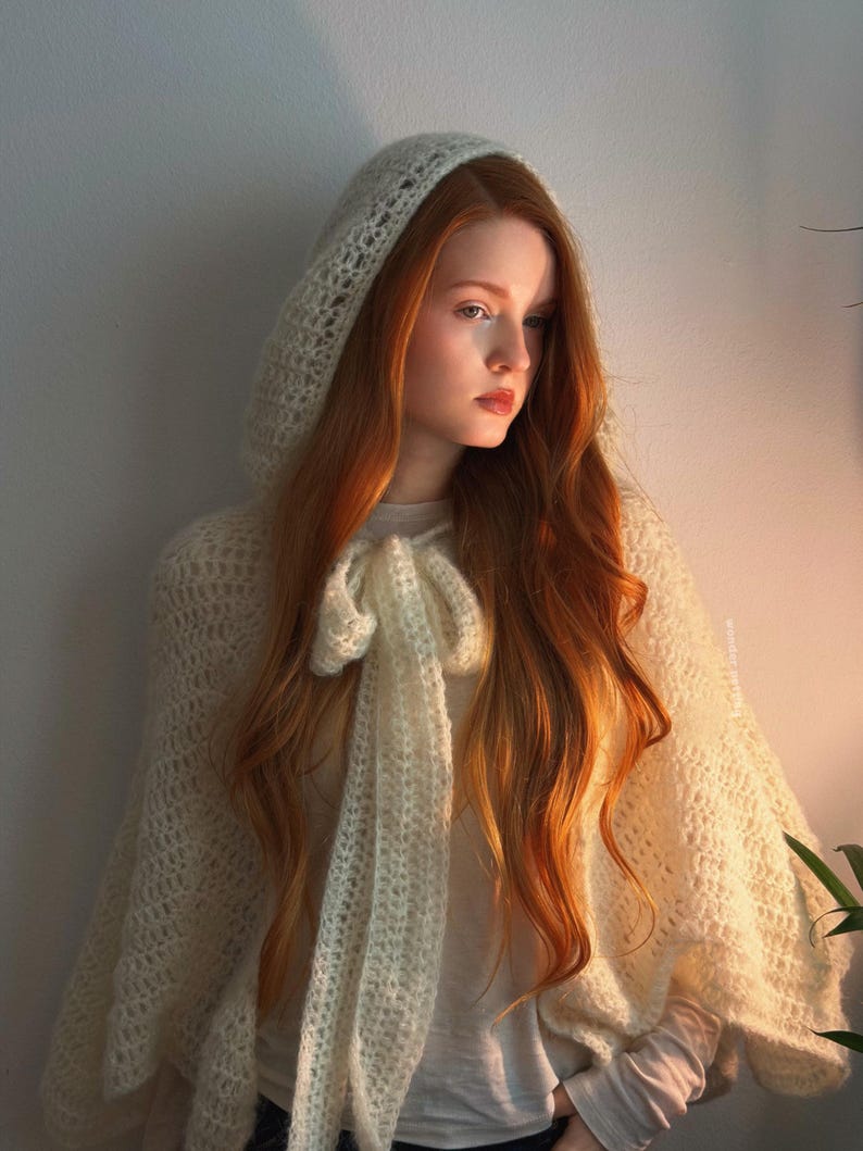 May include: A cream-colored, hooded crochet cape with a bow detail. The cape is worn over a white long-sleeved shirt. The model has long, red hair and is lit by warm sunlight.