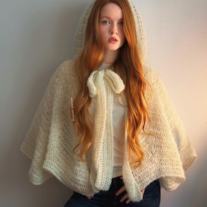 May include: Cream-colored, hooded cape with a tie closure. The cape is made of a loosely woven, textured material. The hood and cape drape over the shoulders. The model is wearing a white shirt and blue jeans.