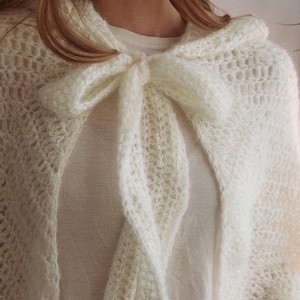 May include: Cream-colored, open-weave knitted cape with a large bow at the neckline. The cape is made of a soft, fluffy yarn and drapes over the shoulders. The bow is a prominent feature, adding a touch of elegance to the design.