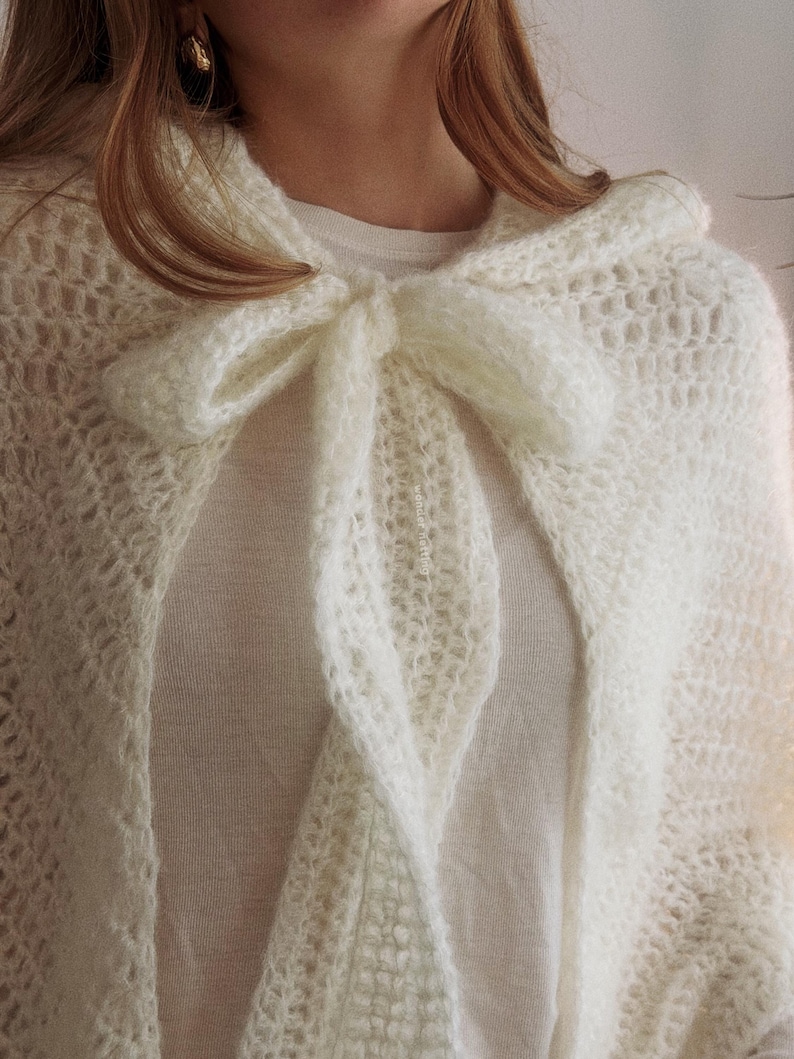 May include: Cream-colored, open-weave knitted cape with a large bow at the neckline. The cape is made of a soft, fluffy yarn and drapes over the shoulders. The bow is a prominent feature, adding a touch of elegance to the design.