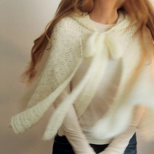 May include: A cream-colored, hooded cape with a bow detail. The cape is made of a soft, open-weave knit fabric. The image shows the cape in motion, with the fabric flowing. The cape is worn over a white long-sleeved shirt and blue jeans.
